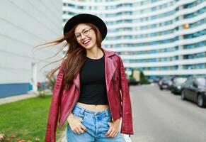 härlig flicka med uppriktig leende Framställ på de gata . blåsigt hårstrån . bär runda glasögon, röd läder jacka. urban bakgrund. foto