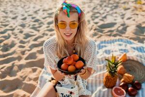 skön blond flicka innehav tallrik med persika och avkopplande på de kväll tropisk strand, solnedgång färger. Sammanträde på handduk. tropisk frukter. bär eleganta pannband och boho klänning. friska liv. foto