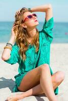 utomhus- sommar porträtt av Söt ung blond skön kvinna i Häftigt solglasögon Framställ på de solig tropisk strand. bär eleganta strand utrusta. foto