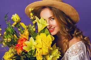 stänga upp vår porträtt av skön blond ung lady i eleganta sugrör sommar hatt innehav färgrik vår blomma bukett nära lila vägg bakgrund. foto