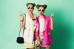 mode studio bild av två ung kvinnor i eleganta tillfällig vår utrusta har roligt, visa tunga. ljus trendig färger, eleganta frisyr med bullar , Häftigt solglasögon. vänner porträtt. foto