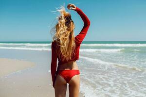 stänga upp utomhus- skott av ung sexig blond kvinna i röd bikini, solbad på hav Strand. hav strand bakgrund. perfekt smal kropp och stånga med sand. högtider och resa begrepp. Foto från tillbaka.