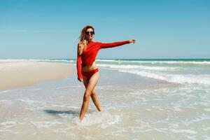 Lycklig blond kvinna i vatten leende Lycklig gående mot kamera. frihet och högtider begrepp. fantastiskt tropisk strand. bär röd bikini. perfekt solbränna kropp och smal figur. foto