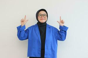 skön ung asiatisk muslim kvinna i glasögon, hijab och bär blå blazer leende självsäker och Lycklig med både händer pekande upp foto