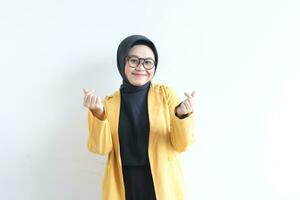 skön ung asiatisk muslim kvinna, bär glasögon och gul blazer gestikulerar koreanska kärlek tecken medan leende foto