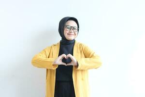 skön ung asiatisk kvinna bär glasögon, hijab och bär gul blazer gestikulerar kärlek tecken med henne hand foto