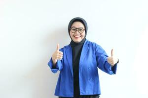 skön ung asiatisk muslim kvinna, bär glasögon och blå blazer med hand gest av godkännande eller ok foto