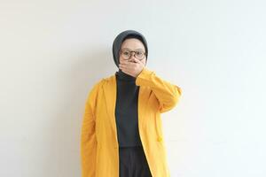 skön ung asiatisk muslim kvinna, bär glasögon och gul blazer med chockade uttryck medan beläggning mun foto