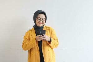 skön ung asiatisk kvinna i glasögon, hijab och bär en gul blazer ser på henne mobil telefon med Lycklig ansikte foto