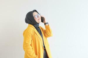 porträtt av skön asiatisk kvinna i hijab, glasögon och bär gul blazer framställning telefon ring upp medan leende foto