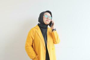 porträtt av skön asiatisk kvinna i hijab, glasögon och bär gul blazer framställning telefon ring upp medan tänkande foto
