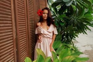 graciös asiatisk kvinna med perfekt hud och hibiskus blomma i hårstrån Framställ över trä vägg och tropisk växter. sommar mode porträtt. foto