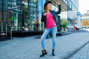 Lycklig blond kvinna Framställ på modern gator, dricka kaffe eller cappuccino. eleganta höst utrusta, läder jacka och stickat Tröja. urban bakgrund. rosa solglasögon. foto