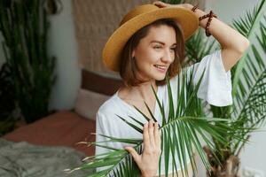 romantisk kvinna med uppriktig leende Sammanträde på säng, njuter solig morgon- i henne eleganta platt i boho stil. foto