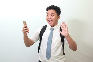 indonesiska senior hög skola studerande bär vit skjorta enhetlig med grå slips vinka hans hand till mobil telefon, ordspråk Hej medan håller på med video ringa upp. isolerat bild på vit bakgrund foto