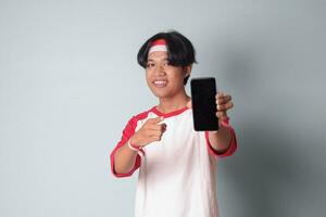porträtt av attraktiv asiatisk man i t-shirt med röd och vit band på huvud, innehav och som visar tom skärm av mobil telefon för mock-up. isolerat bild på grå bakgrund foto