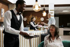 ankomst i hotell reception, äldre manlig kund checkar i med receptionist bistånd på främre skrivbord. caucasian kvinna concierge mottar personlig dokumentera från gammal man för slät registrering. foto