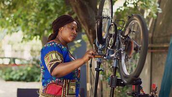 porträtt skott av entusiastisk svart kvinna försiktigt inspekterande för skadestånd på cykel hjul för underhåll. ung afrikansk amerikan kvinna reparation henne egen cykel i gård, kontroll pedaler till fixera. foto