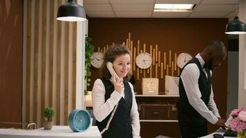 hotell personal svar samtal på reception, använder sig av fast telefon telefon till göra avlägsen bokningar på främre skrivbord. vänlig receptionist förvaring bevis av boende och rum reservationer. foto