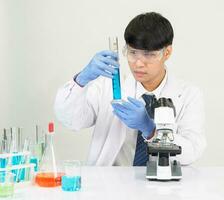 porträtt asiatisk man studerande forskare bär en läkare klänning i de labb ser hand på kemist. orsakade förbi blandning reagens i vetenskaplig forskning laboratorier med testa rör och mikroskop på de tabell foto