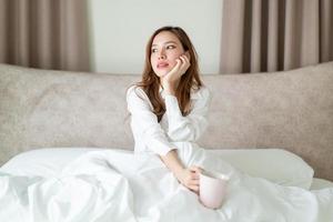 porträtt vacker asiatisk kvinna vaknar och håller kaffekopp eller mugg på sängen på morgonen foto