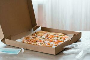 hämtmat mat. pizza i en kartong låda, handskar och skydda mask på de tabell i de kök. foto