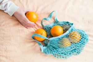 kvinnas hand veck färsk apelsiner i en maska väska på en tyg bakgrund. noll avfall foto