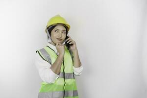 en omtänksam ung kvinna arbetskraft arbetstagare bär säkerhet hjälm och väst medan få en telefon ring upp och hand på henne haka, isolerat förbi vit bakgrund. arbetskraftens dag begrepp. foto
