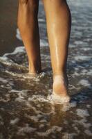 närbild detalj av kvinna ben gående längs de stranden. kvinnors ben och fötter promenad längs de sand och hav vågor, längs de strand. avslappning resa begrepp. vertikal Foto. foto