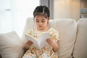 glad Lycklig asiatisk barn bebis flicka leende och läsning bok medan Sammanträde på soffa soffa i levande rum på Hem. flicka relex läsning bok leende på soffa i de hus. tillbaka till skola begrepp. foto