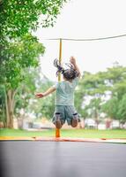 asiatisk barn flicka är Hoppar på trampolin på lekplats bakgrund. Lycklig skrattande unge utomhus i de gård på sommar semester. hoppa hög på trampolin. aktivitet barn i de dagis skola. foto