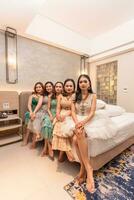 en grupp av asiatisk kvinnor Sammanträde tillsammans på en vit säng medan bär klänningar och smink under en fest på en vänner hus foto
