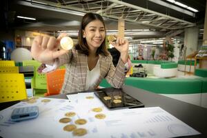 skön asiatisk affärskvinna i närbild skott är visad innehav en bitcoin i henne hand medan leende med en dokumentera synlig på henne skrivbord. foto