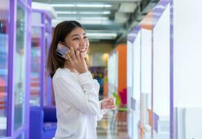 skön asiatisk affärskvinna är stående och talande på de telefon med en Lycklig leende medan kommunicerar med henne kunder, digital marknadsföring. foto