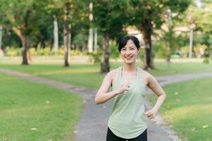 passa ung asiatisk kvinna joggning i parkera leende Lycklig löpning och njuter en friska utomhus- livsstil. kvinna joggare. kondition löpare flicka i offentlig parkera. friska livsstil och wellness varelse begrepp foto