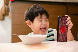 de ansikte av ett asiatisk barn äter medan ser på de telefon foto