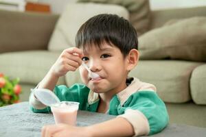 en pojke lyckligt användningar en sked till skopa upp yoghurt. foto