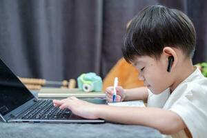 asiatisk pojke lyssnande till audio och skrivning ner läxa medan studerar uppkopplad på bärbar dator. foto