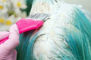 frisör använder sig av rosa borsta medan applicering måla till kvinna kund med smaragd- hår Färg under bearbeta av blekning hår rötter foto