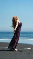 Lycklig kvinna i lång klänning står på strand med huvud ser uppåt, ögon stängd på bakgrund av hav foto