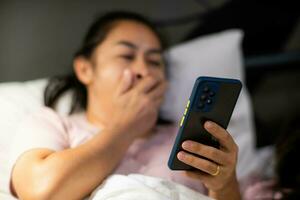 ung asiatisk kvinna liggande på vit säng och spelar smartphone på natt. kvinna i säng med telefon. hälsa och social begrepp. foto