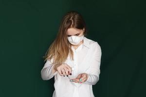 ung blond flicka i en vit blus och en mask använder ett antiseptiskt medel