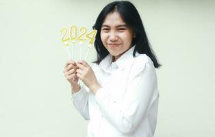 porträtt av Lycklig asiatisk ung kvinna innehav gyllene ljus siffra 2024 till fira ny år eve ha på sig formell vit kostym ser till kamera isolerat över vit bakgrund foto