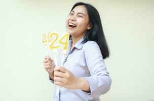 porträtt av upphetsad asiatisk ung kvinna medarbetare skrattande på kamera med stängd öga och visa 2024 figur ljus lyft på hand ha på sig grå formell skjorta, isolerat på vit, ny år eve begrepp foto