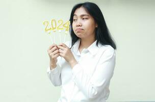 omtänksam asiatisk ung kvinna innehav 2024 figur ljus med nyfikenhet och ser åt sidan till siffra 24 bär vit formell skjorta, isolerat foto