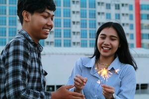 lekfull asiatisk ung par brinnande sparkler med leende ansikte för ny år firande, på urban stad byggnad bakgrund bär flanell och Ränder skjorta tillfällig foto