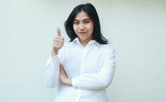 fredlig Framgång asiatisk ung företag kvinna självsäker ger tummen upp med vikta vapen visa godkänna gest, känna nöjd och lugna, bär vit formell kostym skjorta stående över isolerat bakgrund foto