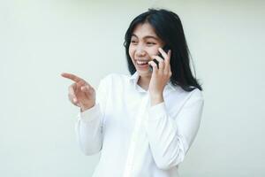 sorglös glädje asiatisk företag kvinna pekande finger och skrattande medan talande på smartphone bär vit kostym formell skjorta, som visar presenter gest, ser bort, stående isolerat bakgrund foto