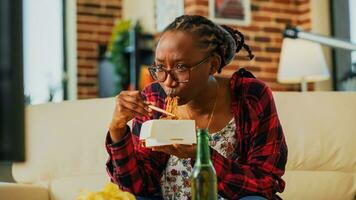 kvinna person använder sig av ätpinnar till äta asiatisk mat, skrattande på favorit filma på tv. ung kvinna äter spaghetti i leverans låda och dricka öl, har roligt på tv. handhållen skott. foto