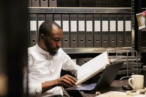 afrikansk amerikan åklagare läsning fil, framställning för fall i polis kontor med arkiv uppgifter. lag tillämpning professionell analyserar forensisk expertis mapp på natt tid foto